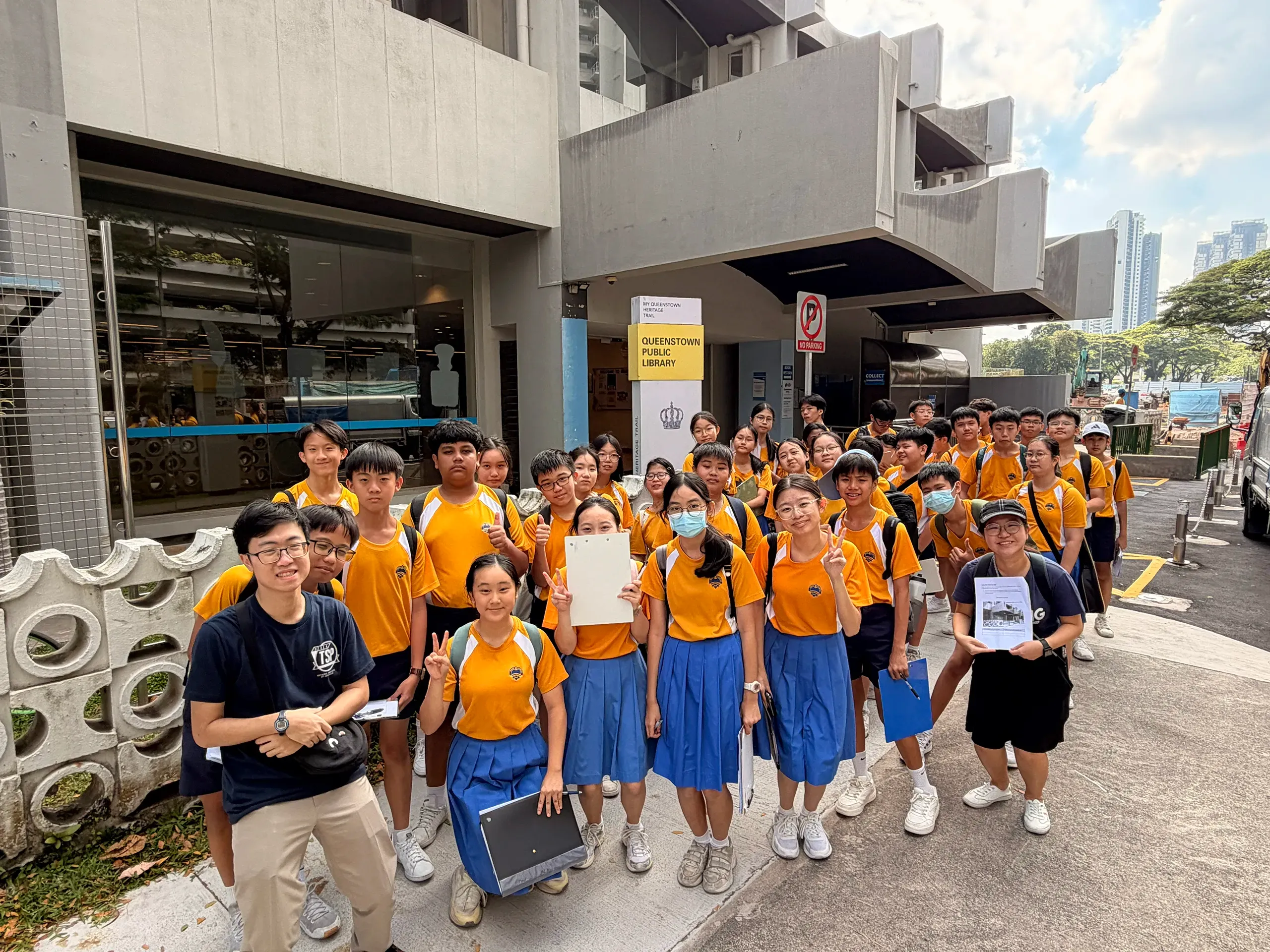 A large group of students in yellow-and-blue school uniforms stands together outside a public library, smiling for a group photo with their teachers. Many hold clipboards, worksheets, and learning materials, indicating participation in a structured educational visit. The library entrance and surrounding urban environment provide the backdrop, highlighting a community-based learning experience that extends beyond the classroom.